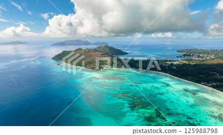 A lush green island stretches along the coast, with colorful coral reefs visible beneath turquoise waters. Praslin, Seychelles. 125988798