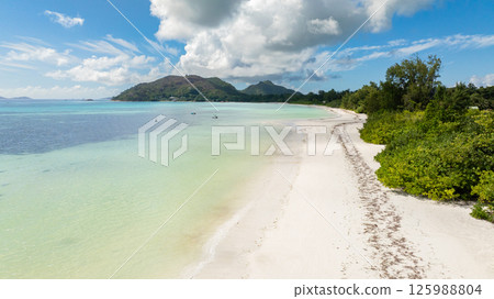 A pristine beach with soft white sand, lush palm trees, and boats floating in turquoise waters near a small island. Praslin, Seychelles. A pristine beach with soft white sand, lush palm trees, and boats floating in turquoise waters near a small island. Praslin, Seychelles. 125988804