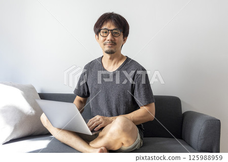 Portrait of mature man wearing spectacles and working from home. Mid adult man sitting on sofa while working on laptop and looking at camera. Businessman using laptop in telecommuting from home. 125988959