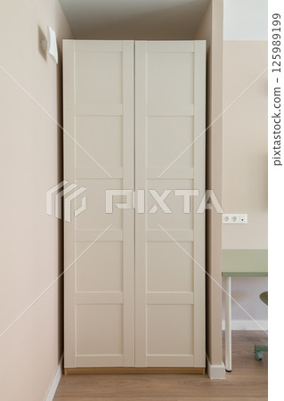 Closed white wardrobe with paneled doors standing in corner next to green desk and wooden floor in interior Closed white wardrobe with paneled doors standing in corner next to green desk and wooden floor in interior 125989199