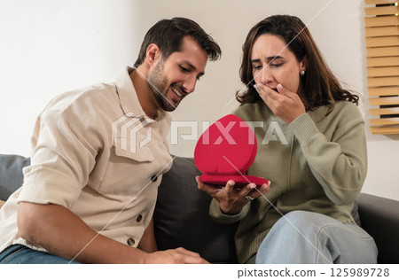 Romantic couple give a surprise gift at home, showing love, relationship, and jewelry in a warm married moment Romantic couple give a surprise gift at home, showing love, relationship, and jewelry in a warm married moment 125989728