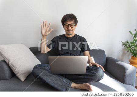 Young smiling asian man relaxing using laptop computer working and video conference meeting at home.Young creative man looking at screen typing message with smartphone.work from home concept 125990200