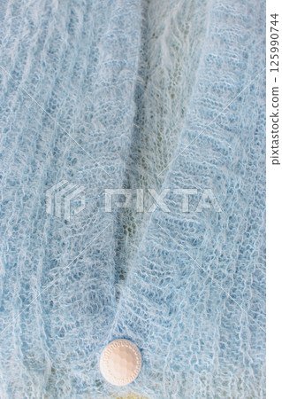 Blue knitted cardigan with white button on white background. Soft blue knitted sweater in details. Knitting pattern background. Summer knitted fashion. Cozy hobby. Craft product concept.  125990744