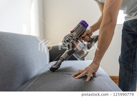 Adult Man in jeans and T-shirt vacuuming a sofa 125990779