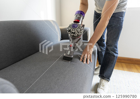 Adult Man in jeans and T-shirt vacuuming a sofa 125990780