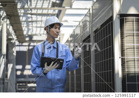 一名身穿工作服的男子正在檢查安裝在建築物屋頂上的戶外空調機組。 125991117