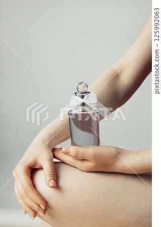 woman hold glass perfume bottle with white bow on neutral background, gentle hand positioning, natural nails, beauty branding, skincare marketing, luxury fragrance, vertical 125992063
