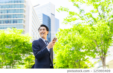 A male business person using a smartphone in a city with fresh greenery 125993409