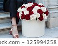 A luxurious bouquet of red and white roses is in a decorative gift box beside a stylishly woman legs in sparkly shoes. Valentines Day, flower store, shop, mock-up, delivery flowers, blog posts 125994258