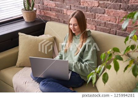 Caucasian young female relaxing on sofa using laptop in cozy brick walled living Caucasian young female relaxing on sofa using laptop in cozy brick walled living 125994281