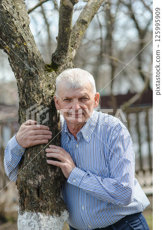 The pensioner is leaning on a tree and smiling beautifully. An elderly man is leaning against a tree. 125995909