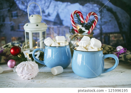 Two cups of hot cocoa with marshmallows and lollipops, lantern with burning candle, winter city landscape in the background Two cups of hot cocoa with marshmallows and lollipops, lantern with burning candle, winter city landscape in the background 125996195