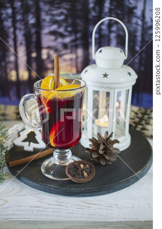 Mulled wine with orange and spices, Christmas decor and burning candles, on a white wooden table against the background of winter nature. 125996208