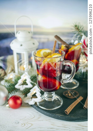 Mulled wine with orange and spices, Christmas decor and burning candles, on a white wooden table against the background of winter nature. Mulled wine with orange and spices, Christmas decor and burning candles, on a white wooden table against the background of winter nature. 125996210