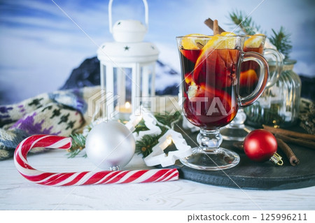Mulled wine with orange and spices, Christmas decor and burning candles, on a white wooden table against the background of winter nature. Mulled wine with orange and spices, Christmas decor and burning candles, on a white wooden table against the background of winter nature. 125996211