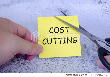 Hand holding a yellow sticky note with the words Cost Cutting written on it, while a pair of scissors is cutting through the note. Hand holding a yellow sticky note with the words Cost Cutting written on it, while a pair of scissors is cutting through the note. 125996757