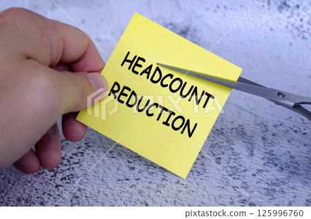 Hand holding a yellow sticky note with the words Headcount Reduction written on it, while a pair of scissors is cutting through the note. Hand holding a yellow sticky note with the words Headcount Reduction written on it, while a pair of scissors is cutting through the note. 125996760