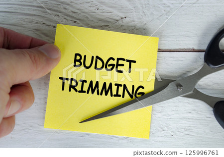 Hand holding a yellow sticky note with the words Budget Trimming written on it, while a pair of scissors is cutting through the note 125996761