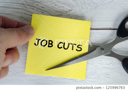 Hand holding a yellow sticky note with the words Job Cuts written on it, while a pair of scissors is cutting through the note. 125996763