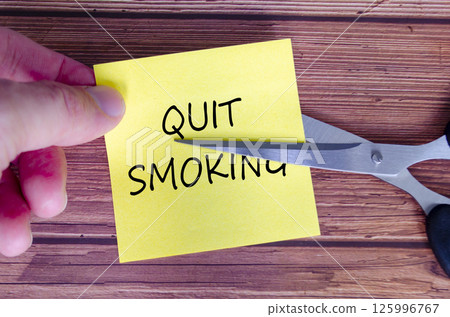 Hand holding a yellow sticky note with the words Quit Smoking written on it, while a pair of scissors is cutting through the note. Hand holding a yellow sticky note with the words Quit Smoking written on it, while a pair of scissors is cutting through the note. 125996767