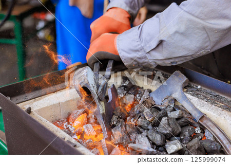 Craftsman works diligently at forge, shaping molten metal to axe while surrounded by glowing coals, tools 125999074