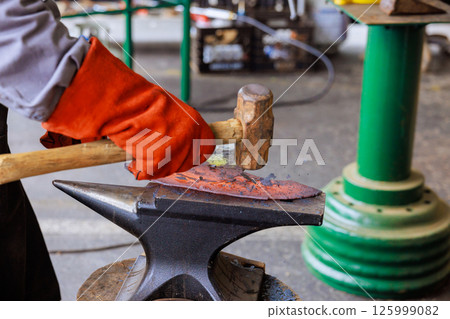 Craftsman shapes metal on an anvil using hammer in during axe making workshop setting. 125999082