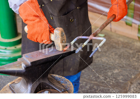 Blacksmith uses hammer to shape heated metal into garden fork while wearing protective gloves. 125999083
