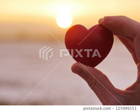 A hand holding a red heart in front of a sunset A hand holding a red heart in front of a sunset 125999151