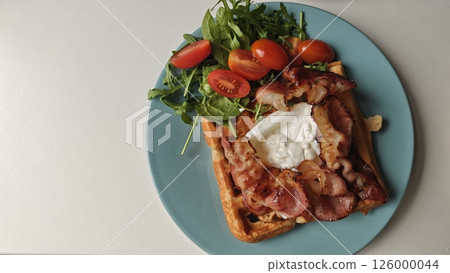 Savory waffle breakfast with crispy bacon, poached egg, cherry tomatoes and arugula on blue plate. Concept of protein-rich brunch, healthy eating and modern food styling Savory waffle breakfast with crispy bacon, poached egg, cherry tomatoes and arugula on blue plate. Concept of protein-rich brunch, healthy eating and modern food styling 126000044