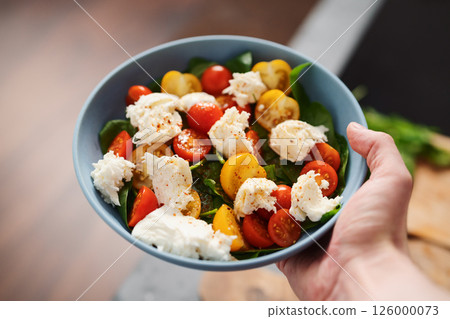 amn hand hold salad with red and yellow cherry tomatoes , mozzarella and spinach in blue bowl 126000073