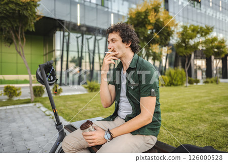 Man with curly hair sits on a park bench, smokes a cigar and rests 126000528
