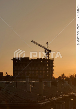 sunset casting shadows over construction site with crane towering above rooftops in city landscape. 126000730