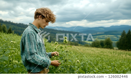 A young farmer examines plants in a scenic field with rolling hills and cloudy skies 126002446