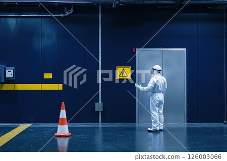 Industrial safety protocol: worker in protective gear in a secure facility with warning signs and caution cone 126003066