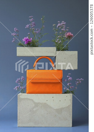 Vibrant orange handbag in minimalist floral display on concrete blocks for chic fashion branding 126005378