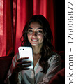 Young woman smiling in cozy setting using smartphone against red curtains - technology and relaxation 126006872