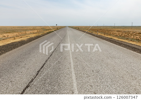 Straight asphalt road stretches through arid landscape under cloudy sky Atyrau Region Kazakhstan 126007347