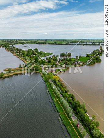 Aerial view of a scenic lakeside village with narrow peninsulas, green fields, and houses surrounded by calm waters under a cloudy sky in the Dutch countryside. Aerial view of a scenic lakeside village with narrow peninsulas, green fields, and houses surrounded by calm waters under a cloudy sky in the Dutch countryside. 126008371