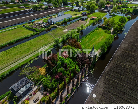 Aerial view of a lush countryside garden surrounded by canals and farmlands, with a cozy house and colorful trees, set in a peaceful rural landscape under a clear blue sky. Aerial view of a lush countryside garden surrounded by canals and farmlands, with a cozy house and colorful trees, set in a peaceful rural landscape under a clear blue sky. 126008372