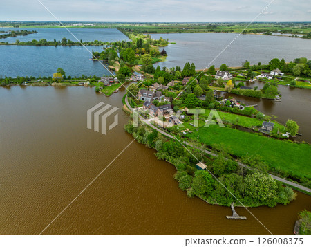 Aerial view of a scenic lakeside village with narrow peninsulas, green fields, and houses surrounded by calm waters under a cloudy sky in the Dutch countryside. Aerial view of a scenic lakeside village with narrow peninsulas, green fields, and houses surrounded by calm waters under a cloudy sky in the Dutch countryside. 126008375