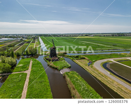 Aerial view of a Dutch countryside with a traditional windmill by a canal, green fields, and a nearby road with moving cars under a clear blue sky. Aerial view of a Dutch countryside with a traditional windmill by a canal, green fields, and a nearby road with moving cars under a clear blue sky. 126008376