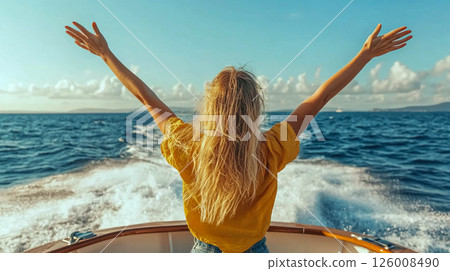 A girl from the back standing on board a moving yacht with her arms raised. The seascape stretches beyond it. 126008490