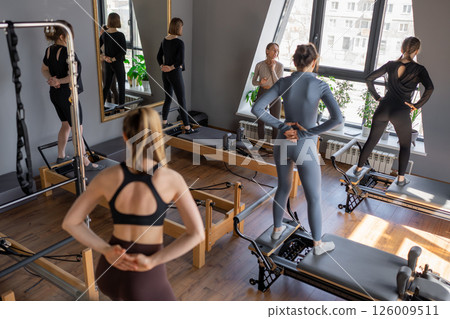 Group of caucasian women exercising with trainer on pilates reformers in a bright studio. Their workout focuses on core strength and flexibility in a modern fitness environment 126009511