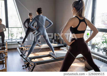 Young caucasian women exercising on pilates reformers in a bright studio. Their workout focuses on core strength and flexibility in a modern fitness environment. Young caucasian women exercising on pilates reformers in a bright studio. Their workout focuses on core strength and flexibility in a modern fitness environment. 126009515
