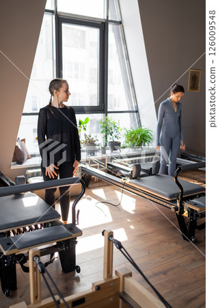 Young caucasian woman doing roll down exercise in a pilates bright studio. Their workout focuses on core strength and flexibility in a modern fitness environment. Young caucasian woman doing roll down exercise in a pilates bright studio. Their workout focuses on core strength and flexibility in a modern fitness environment. 126009548