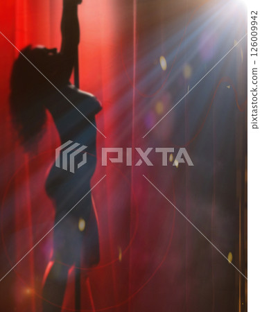 Silhouette of exotic dancer with long curly hair standing on pole in front of orange lights, backlit and cinematic. 126009942