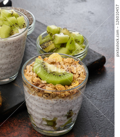 Two glasses of chia seed pudding, one topped with diced kiwi and the other with granola and banana chips, are on a dark surface with ingredients Two glasses of chia seed pudding, one topped with diced kiwi and the other with granola and banana chips, are on a dark surface with ingredients 126010457