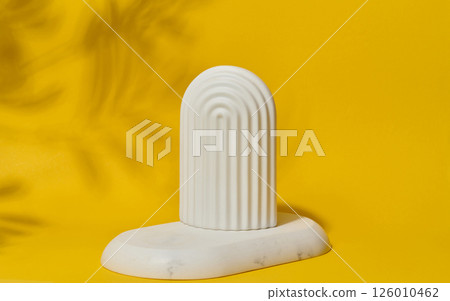 Small, white ceramic decorative arch with vertical ribbed texture stands on a bright yellow surface with blurred leaf shadows. Small, white ceramic decorative arch with vertical ribbed texture stands on a bright yellow surface with blurred leaf shadows. 126010462