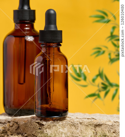 Amber glass dropper bottles on a natural stone surface with a yellow background and green plant elements. Amber glass dropper bottles on a natural stone surface with a yellow background and green plant elements. 126010480