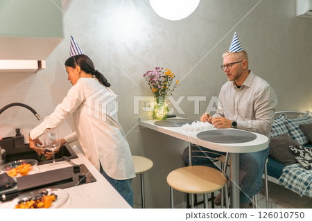 Birthday Party Couple Kitchen: Celebrating intimate birthday at home; woman prepares food, man sits; cozy atmosphere. 126010750